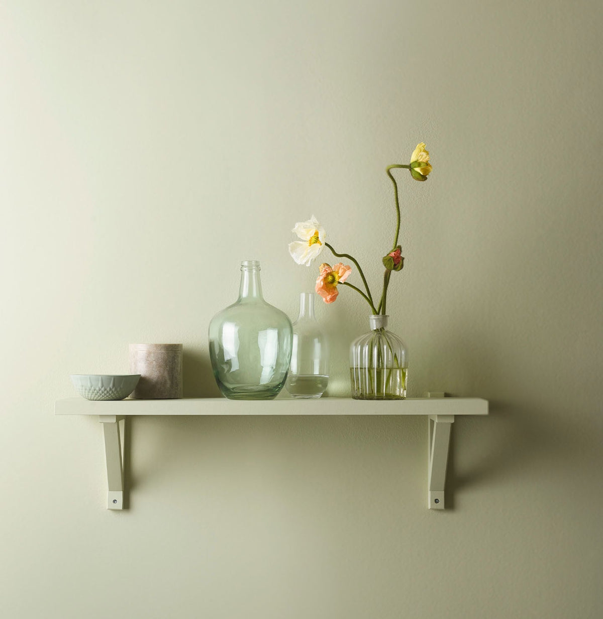 Hylle på veggen innredet med vakre glassvaser og blomster, mot en gulgrønn vegg malt med Jotun 8222 Jurrasic Grey for et friskt og lett uttrykk.