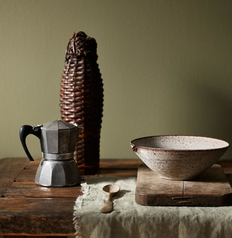 Rustic wooden table with a metal espresso maker and ceramic bowl with a olive green background painted with the Jotun colour 8597 Seaweed Green.