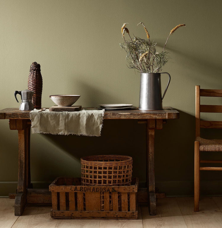 Rustic wooden table with vintage kitchenware and a wicker basket against a olivegreen wall painted with the colour 8597 Seaweed Green from Jotun.