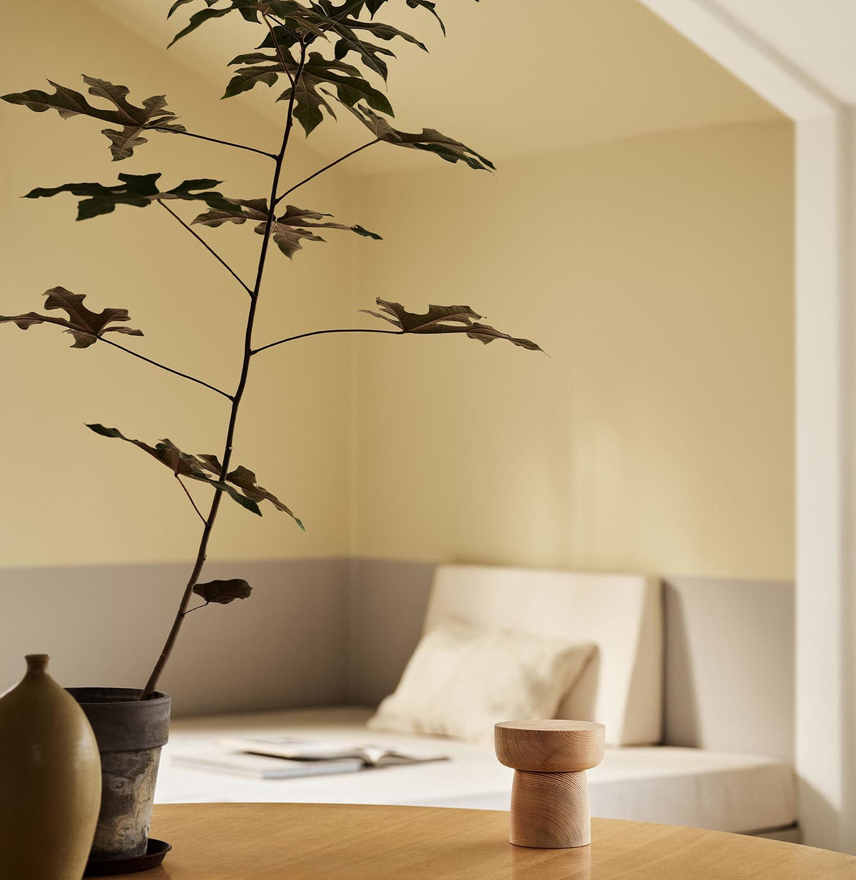 A potted plant and a daybed with light bedding against muted, greenish yellow walls painted in the colour 1965 Ginger Tea from Jotun.