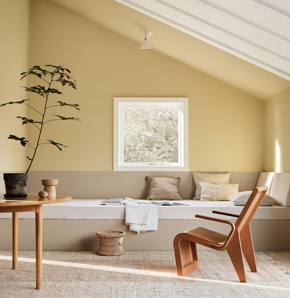 A daybed, wodden chair and a potted plant against muted, greenish yellow walls painted in the colour 1965 Ginger Tea from Jotun. Daybed painted in the colour 12300 Hazelnut Beige. 