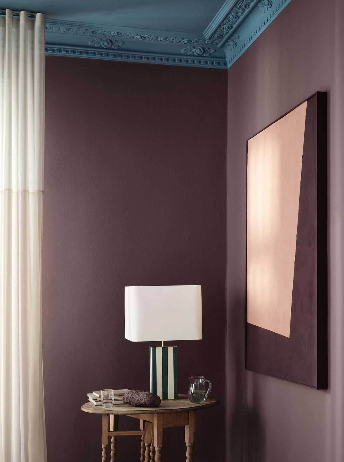 Et hjørne af stuen med rødlilla vægge i Jotun 3343 Dusty Purple, loft malet i blå Jotun 4223 Sama, en stribet lampe på et træbord og et stort abstrakt maleri.